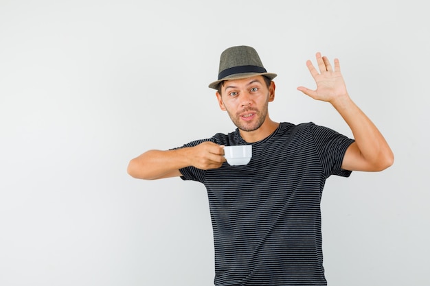 Jovem homem com chapéu de camiseta segurando um copo de bebida, acenando com a mão e olhando feliz