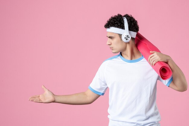 Jovem homem com roupas esportivas e tapete de ioga na parede rosa