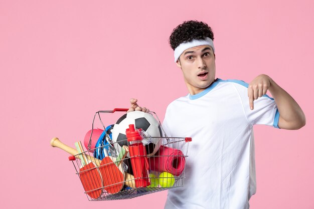 Jovem homem com roupas esportivas e uma cesta cheia de coisas esportivas na parede rosa