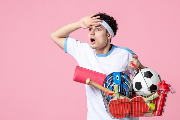 Jovem homem com roupas esportivas e uma cesta cheia de coisas esportivas na parede rosa