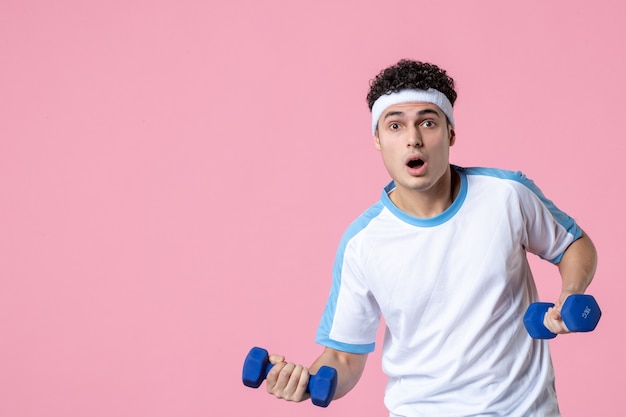 Jovem homem com roupas esportivas, malhando de frente com halteres na parede rosa