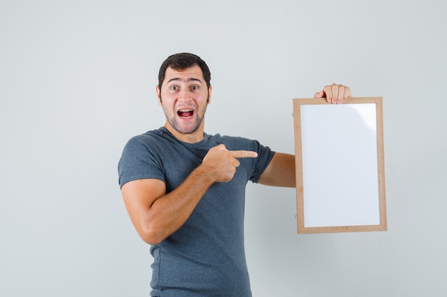 Foto grátis jovem homem com uma camiseta cinza apontando para uma moldura vazia e parecendo feliz