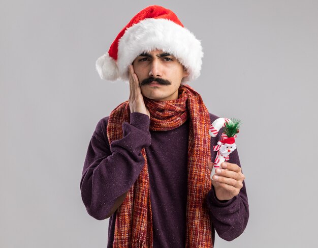 Jovem homem de bigode usando chapéu de Papai Noel com lenço quente no pescoço segurando uma bengala de doces de Natal e parecendo confuso