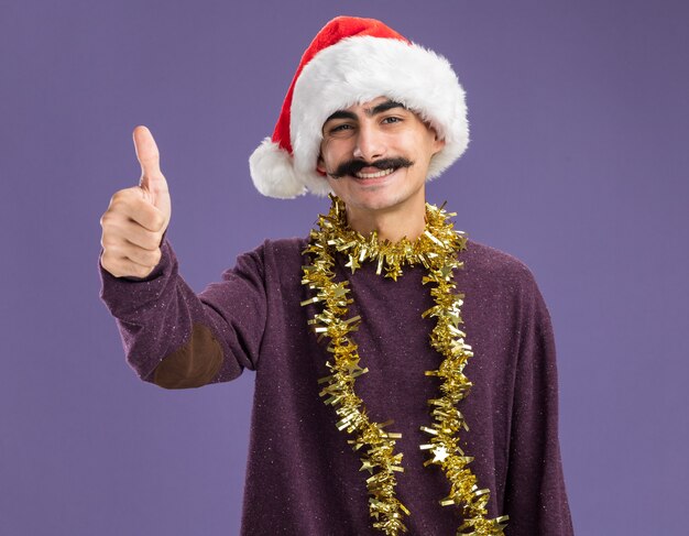 Jovem homem de bigode usando chapéu de Papai Noel de Natal com enfeites em volta do pescoço olhando para a câmera com um sorriso no rosto mostrando os polegares em pé sobre um fundo roxo