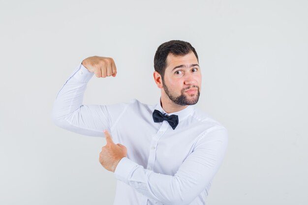 Jovem homem de camisa branca, apontando para seus músculos e parecendo confiante, vista frontal.