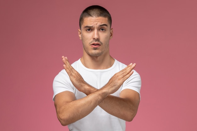 Jovem homem de camisa branca, vista frontal, mostrando sinal de proibição no fundo rosa