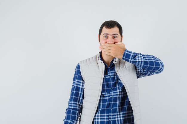 Jovem homem de camisa, jaqueta sem mangas, segurando as mãos na boca e parecendo calmo, vista frontal.