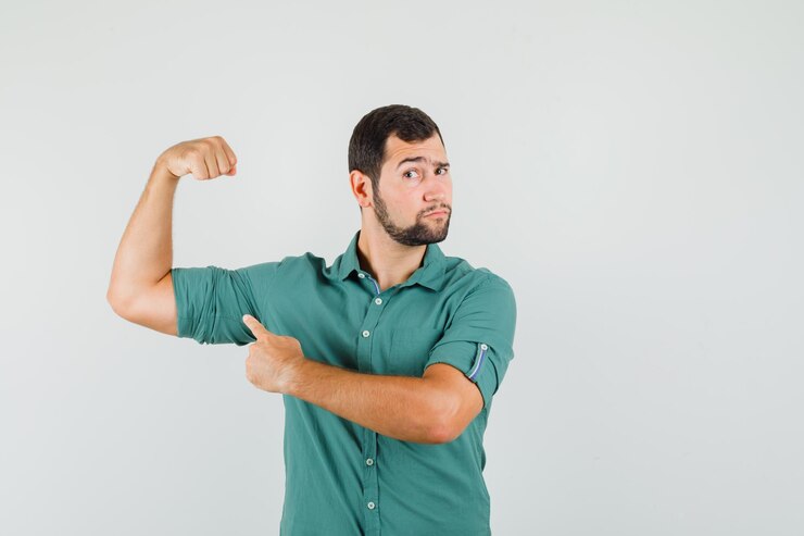 Foto grátis jovem homem de camisa verde, apontando para os músculos do braço e parecendo poderoso, vista frontal.