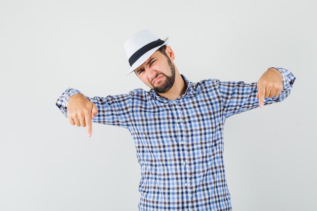 Jovem homem de camisa xadrez, chapéu apontando os dedos para baixo e parecendo descontente, vista frontal.