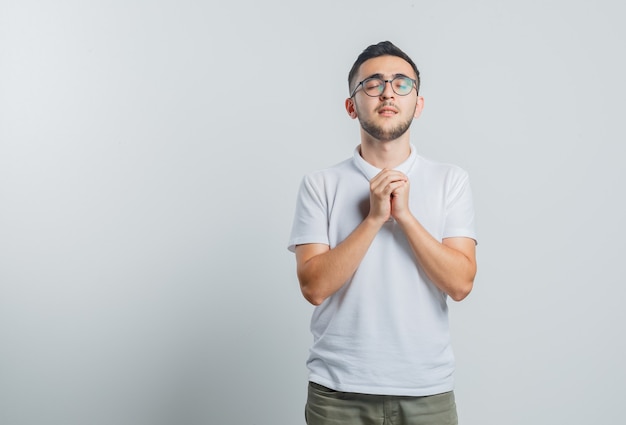 Jovem homem de camiseta branca, calça dando as mãos em gesto de oração e parecendo esperançoso