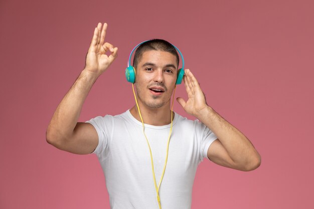 Jovem homem de camiseta branca, de frente, ouvindo música com seus fones de ouvido no fundo rosa claro