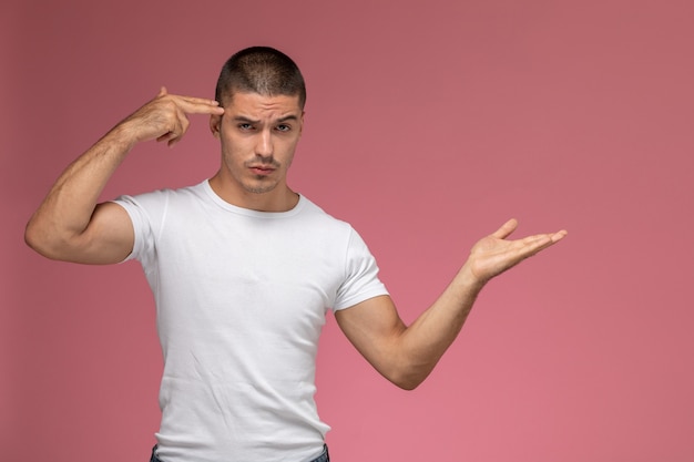 Jovem homem de camiseta branca, de frente, posando e apontando para sua têmpora em um fundo rosa