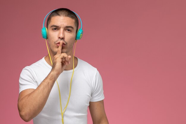 Jovem homem de camiseta branca ouvindo música e mostrando o sinal de silêncio em fundo rosa claro de frente