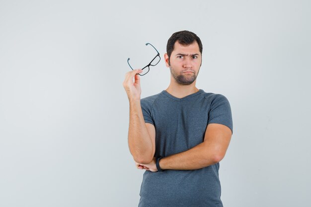 Jovem homem de camiseta cinza segurando óculos e parecendo hesitante