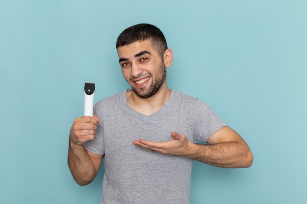 Jovem homem de camiseta cinza segurando um barbeador elétrico com um sorriso na barba de barbear azul