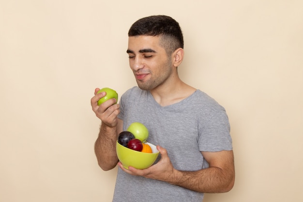 Jovem homem de camiseta cinza segurando um prato com frutas comendo maçã em bege