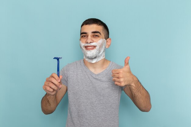 Jovem homem de camiseta cinza segurando uma lâmina de barbear de frente para o homem de barba azul-gelo com espuma de barbear