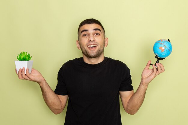 Jovem homem de camiseta preta segurando uma plantinha verde e um globo terrestre com uma expressão encantada no verde