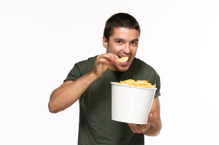 Foto grátis jovem homem de camiseta verde segurando a cesta com batata cip e comendo na parede branca filme prazer solitário