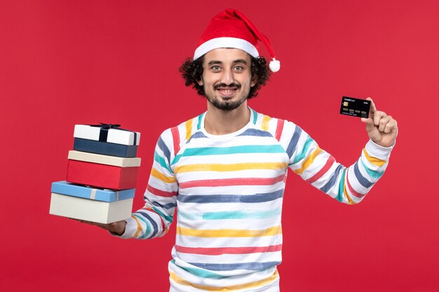Jovem homem de frente com presentes de feriado e cartão do banco na parede vermelha dinheiro de ano novo vermelho