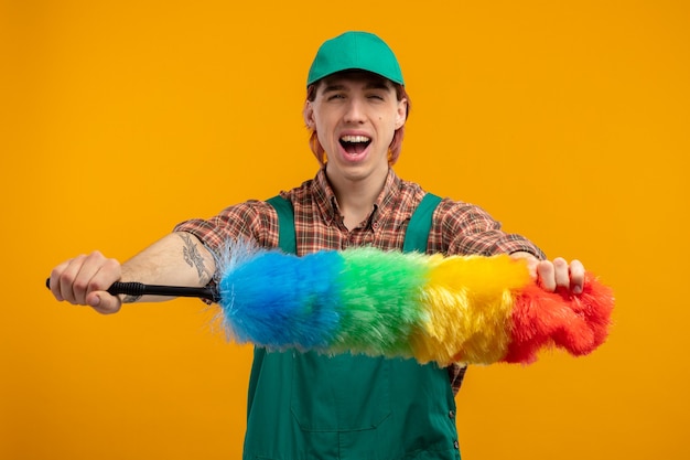 Jovem homem de limpeza com macacão de camisa xadrez e boné segurando um espanador colorido, feliz e animado em pé sobre a parede laranja