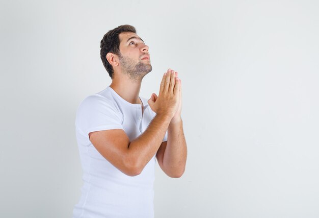 Jovem homem de mãos dadas em gesto de oração em uma camiseta branca e parecendo esperançoso