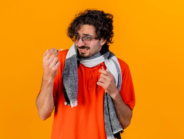 Jovem homem doente, caucasiano, assustado, usando óculos e lenço, segurando uma seringa e uma ampola, olhando para uma seringa isolada na parede laranja com espaço de cópia