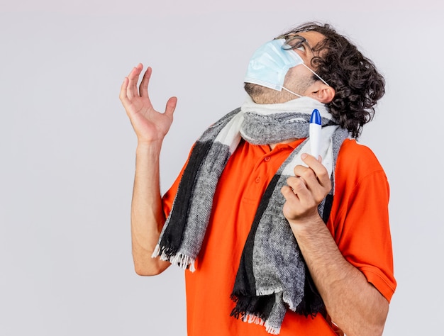 Jovem homem doente, caucasiano, usando óculos e máscara com lenço segurando o termômetro, olhando para cima, mantendo a mão no ar isolado no fundo branco com espaço de cópia