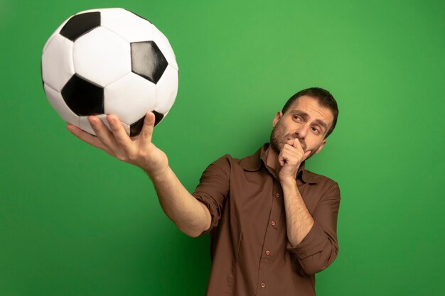 Foto grátis jovem homem duvidoso, caucasiano, estendendo a bola de futebol em direção à câmera, olhando para ela, colocando a mão no queixo isolado em um fundo verde com espaço de cópia