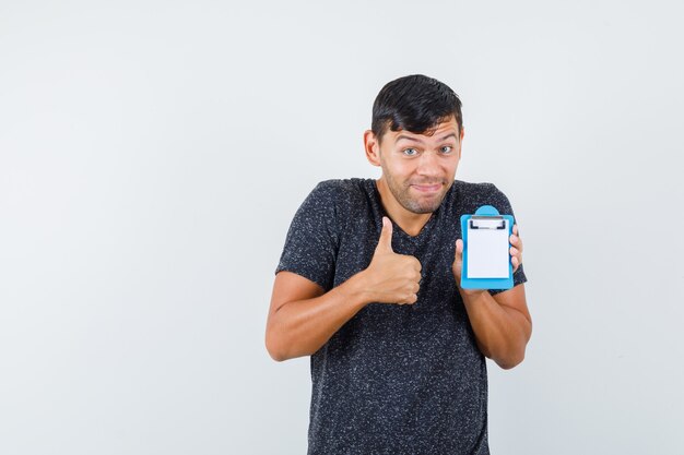 Jovem homem em t-shirt preta aparecendo baque enquanto segura seu pequeno caderno e parece feliz, vista frontal.