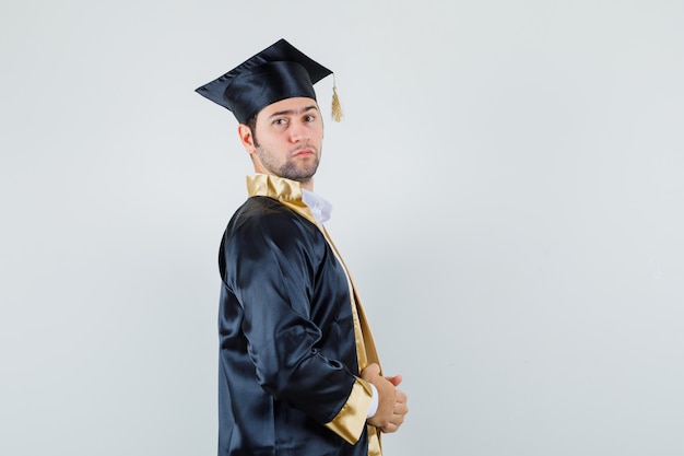 Foto grátis jovem homem em uniforme de pós-graduação segurando seu vestido e parecendo confiante.