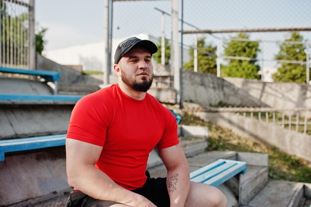 Jovem homem musculoso barbudo brutal usa shorts de camisa vermelha e boné no estádio