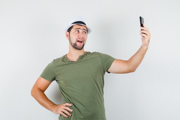 Jovem homem posando enquanto tira uma selfie com camiseta e chapéu verdes e parece alegre