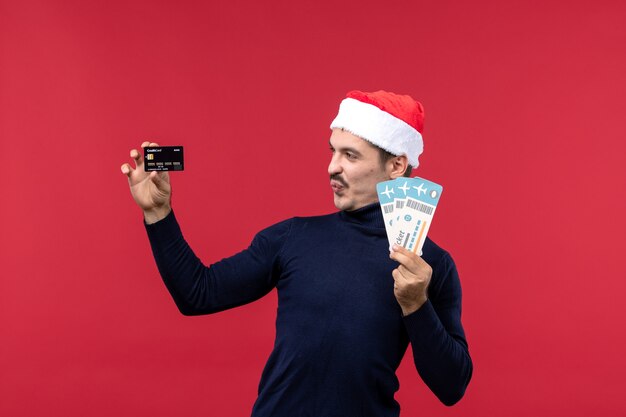 Jovem homem segurando o cartão do banco e os ingressos em fundo vermelho.