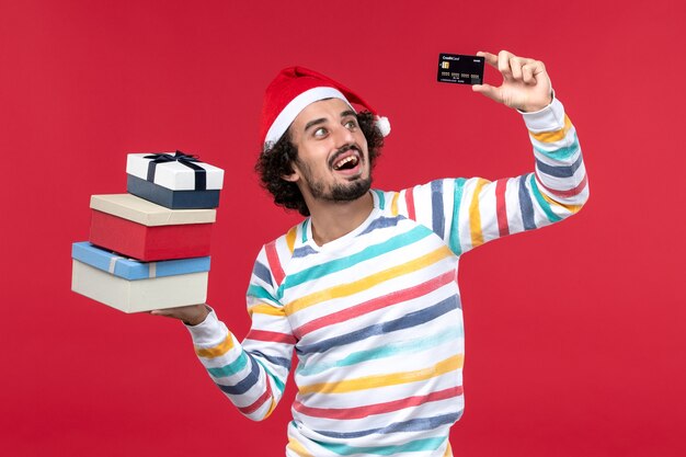 Jovem homem segurando o cartão do banco e presentes na parede vermelha dinheiro de ano novo homem vermelho