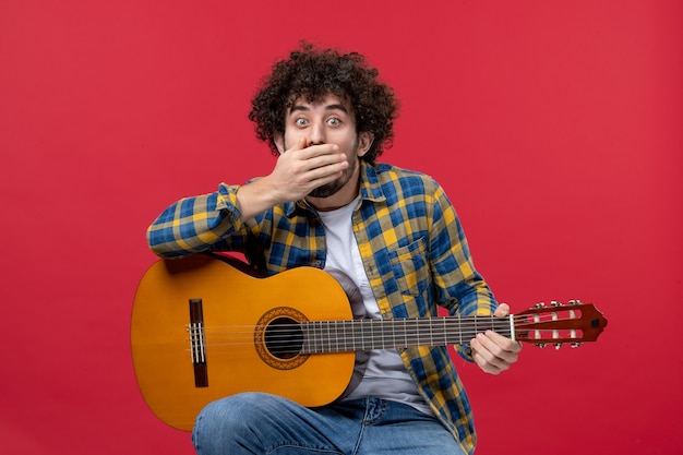 Jovem homem sentado com a guitarra na parede vermelha, vista frontal, aplausos ao vivo músico toca música de concerto de banda colorida