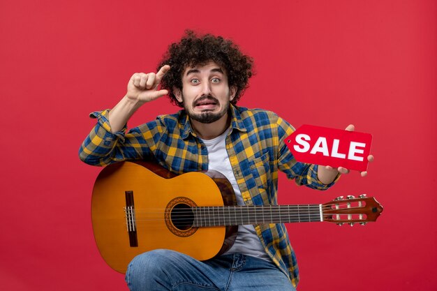 Jovem homem sentado com a guitarra na parede vermelha, vista frontal, música cor, aplausos, concerto de músico ao vivo