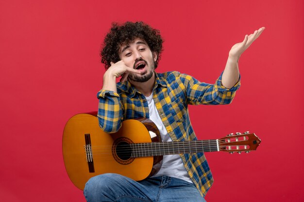 Jovem homem sentado com a guitarra na parede vermelha.