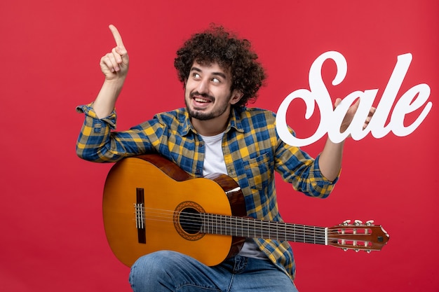 Jovem homem sentado com guitarra na parede vermelha concerto de música de frente aplausos músico ao vivo venda de cores