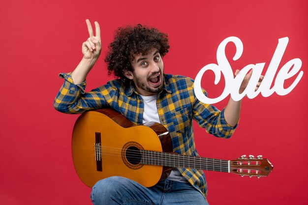 Jovem homem sentado com guitarra na parede vermelha venda de música na parede vermelha tocar concerto aplausos cores ao vivo