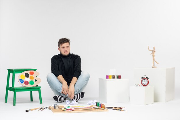Jovem homem sentado em frente a tintas e borlas para desenhar na parede branca desenhar pinturas coloridas.