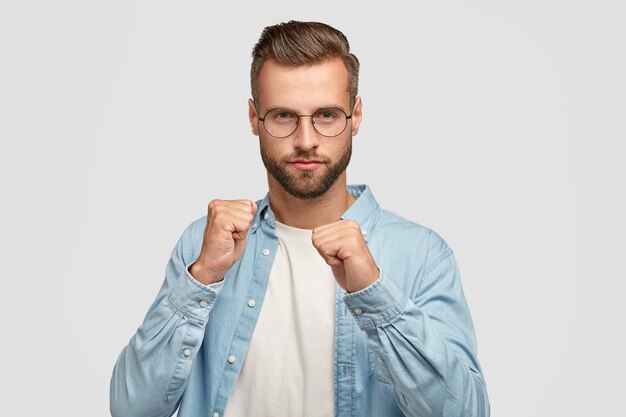 Jovem homem sério com a barba por fazer mostra os punhos, pronto para se defender, usa elegante camisa azul, óculos, posa contra a parede branca. Homem barbudo confiante luta com alguém. Força masculina