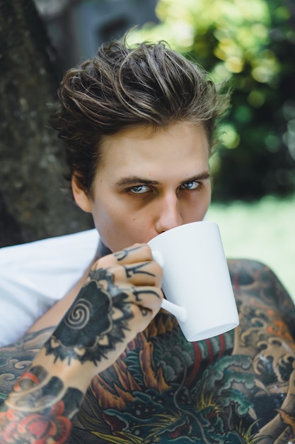 Jovem homem tatuado bonito tomando café da manhã na cama ao ar livre ao ar livre no jardim