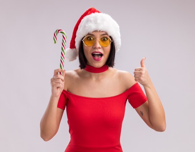 Jovem impressionada com chapéu de Papai Noel e óculos segurando uma bengala de doces de natal, olhando para a câmera mostrando o polegar isolado no fundo branco