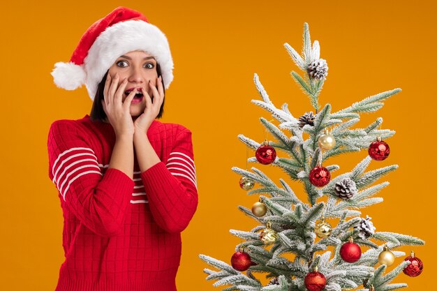Jovem impressionada com chapéu de Papai Noel em pé perto da árvore de Natal decorada, mantendo as mãos no rosto isoladas na parede laranja