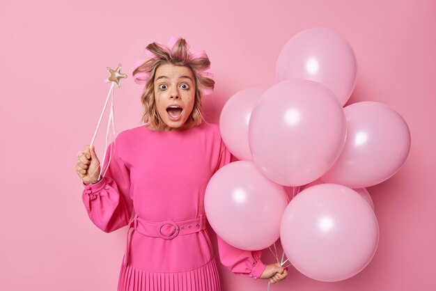 Jovem impressionada usa vestido festivo aplica rolos de cabelo vazou maquiagem depois que cryig segura a varinha mágica e um monte de balões inflados reage em algo incrível isolado sobre a parede rosa