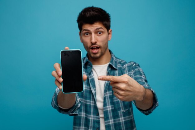 Jovem impressionado esticando o celular em direção à câmera apontando para ele olhando para a câmera isolada em fundo azul