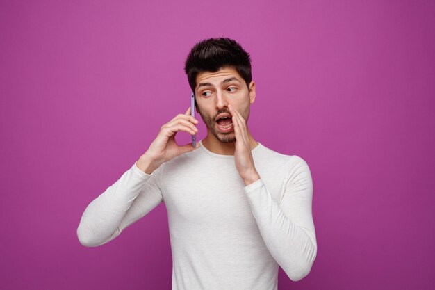 Jovem impressionado falando ao telefone olhando para o lado mantendo a mão perto da boca sussurrando isolado no fundo roxo