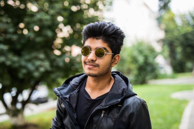 Jovem indiano bonito usando óculos escuros enquanto pensava contra a rua ao ar livre