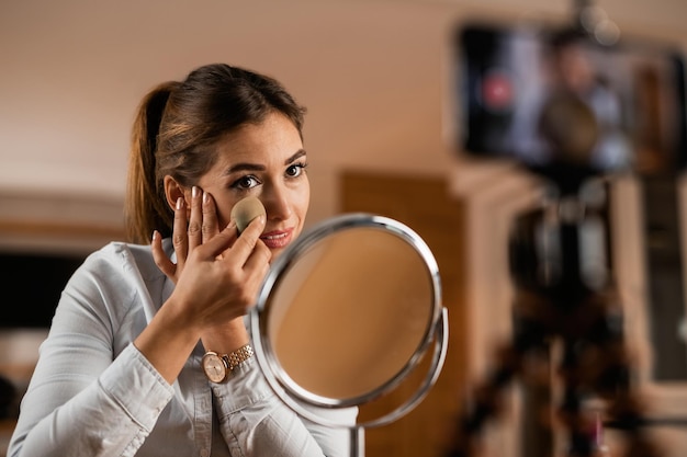 Jovem influenciadora aplicando pó facial enquanto vlog sobre produtos de maquiagem em casa